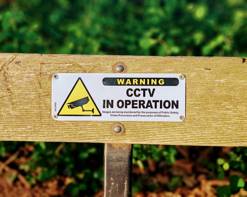 A rectangular metal sign mounted on a horizontal wooden beam, indicating that CCTV is in operation. The sign features a yellow triangle with a black CCTV camera icon on the left side, and bold black a