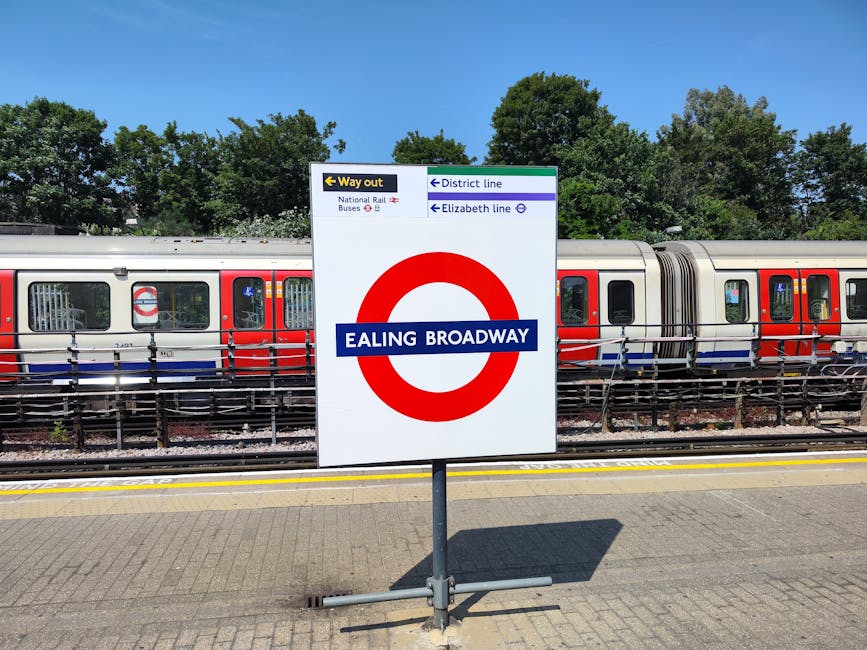 A large white signboard displaying Ealing Broadway station name with a red London Underground roundel, situated on a paved platform. Behind the sign, a modern train with a silver and red exterior is parked on the railway tracks, with several visible windows and doors. To the left of the station name, there is a smaller black and yellow sign indicating the way out, and above it, a blue and white directional sign shows the district and Elizabeth line routes, along with icons for national rail and bus services. The background features lush green trees under a clear blue sky, and the platform edge marked with yellow safety lines. This scene captures the station environment where a home relocation or furniture transport project by Man and a Van Ealing could coordinate loading and moving procedures, with the train serving as part of the transport logistics during house removals.