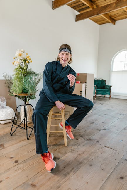 A young man with long hair wearing a dark blue hoodie, dark trousers, and bright orange sports shoes, is sitting on a small wooden stool inside a well-lit room with white walls and a wooden floor. He is smiling and gesturing with his right hand while his left hand rests on his knee. Behind him, there is a side table with a bouquet of white and yellow flowers in a glass vase, and a stack of wrapped cardboard boxes. To the right, an upholstered armchair with a greenish covering is placed near a window with an arched top. The room features exposed wooden ceiling beams, and the scene captures a moment during a home relocation process involving packing and preparing items for transport, reflective of house removal services like those offered by [COMPANY_NAME] in the context of narrow access move tips for Pitshanger Lane, EALING.