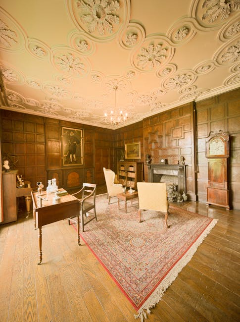 An interior of a room with dark wooden paneled walls and a decorative, ornate ceiling with intricate plasterwork and a central chandelier. On the left side, there is a wooden table with porcelain deco